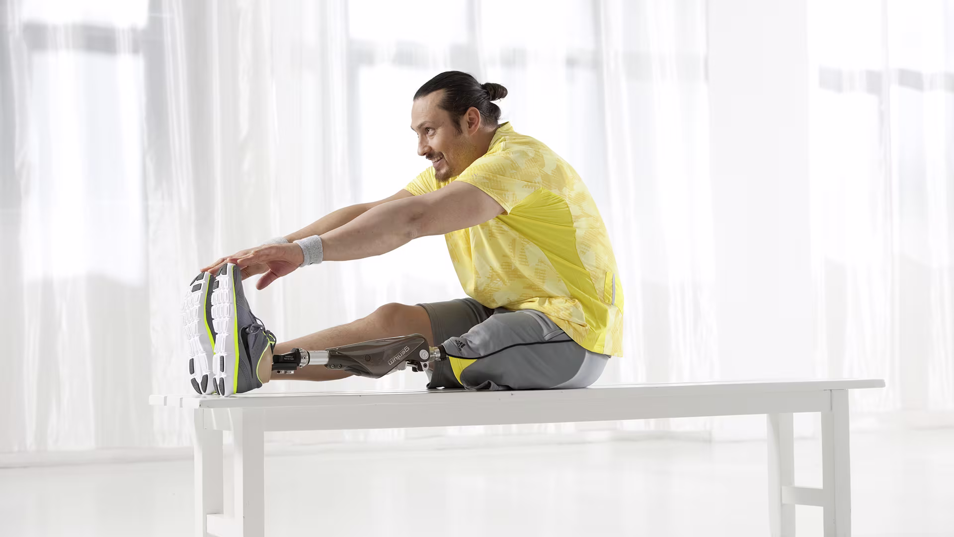 Ein Mann in Trainingskleidung und mit Ottobock Prothese auf einer weißen Bank vor weißem Hintergrund streckt sich und berührt mit den Fingerspitzen die Spitze seiner Schuhe.