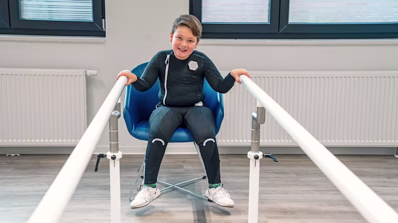 Ein Junge sitz auf einem Stuhl und trägt den exopulse suit von Ottobock.