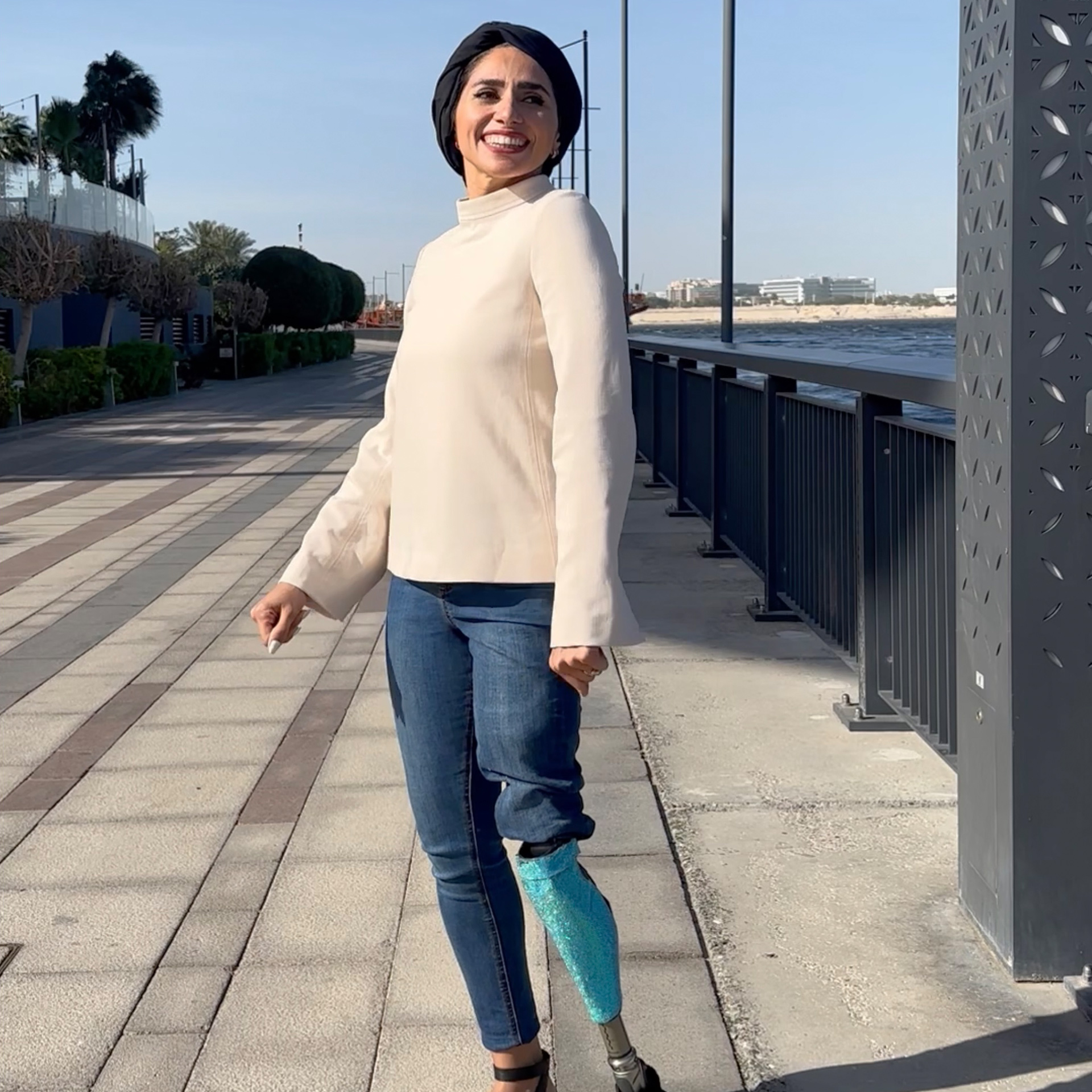 Woman with a lower limb loss Zainab Al-Eqabi with an Ottobock prosthesis stands smiling on a boardwalk by the seashore.