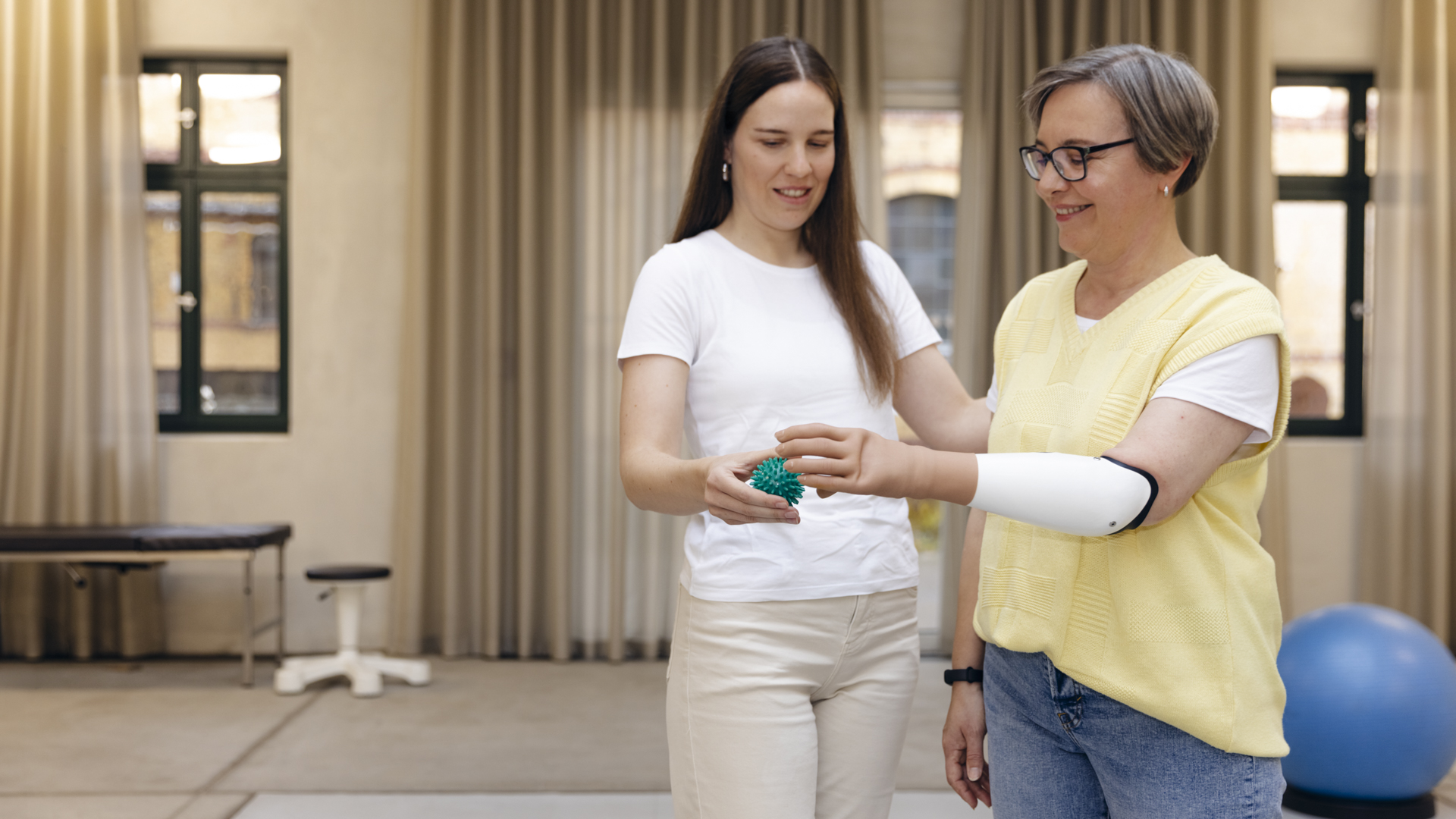 Eine Therapeutin trainiert mit einer armamputierten Frau mit Hilfe eines Gummiballs. Die Frau trägt die speedhand Armprothese von Ottobock.