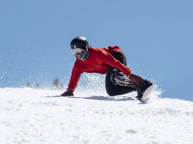 Man in red ski jacket taking an aggressive turn on a snowboard with his hand in the snow