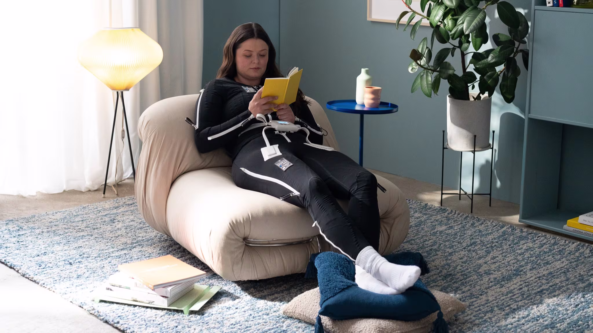 Exopulse Mollii Suit user Lena is relaxing on a sofa chair reading a book. 