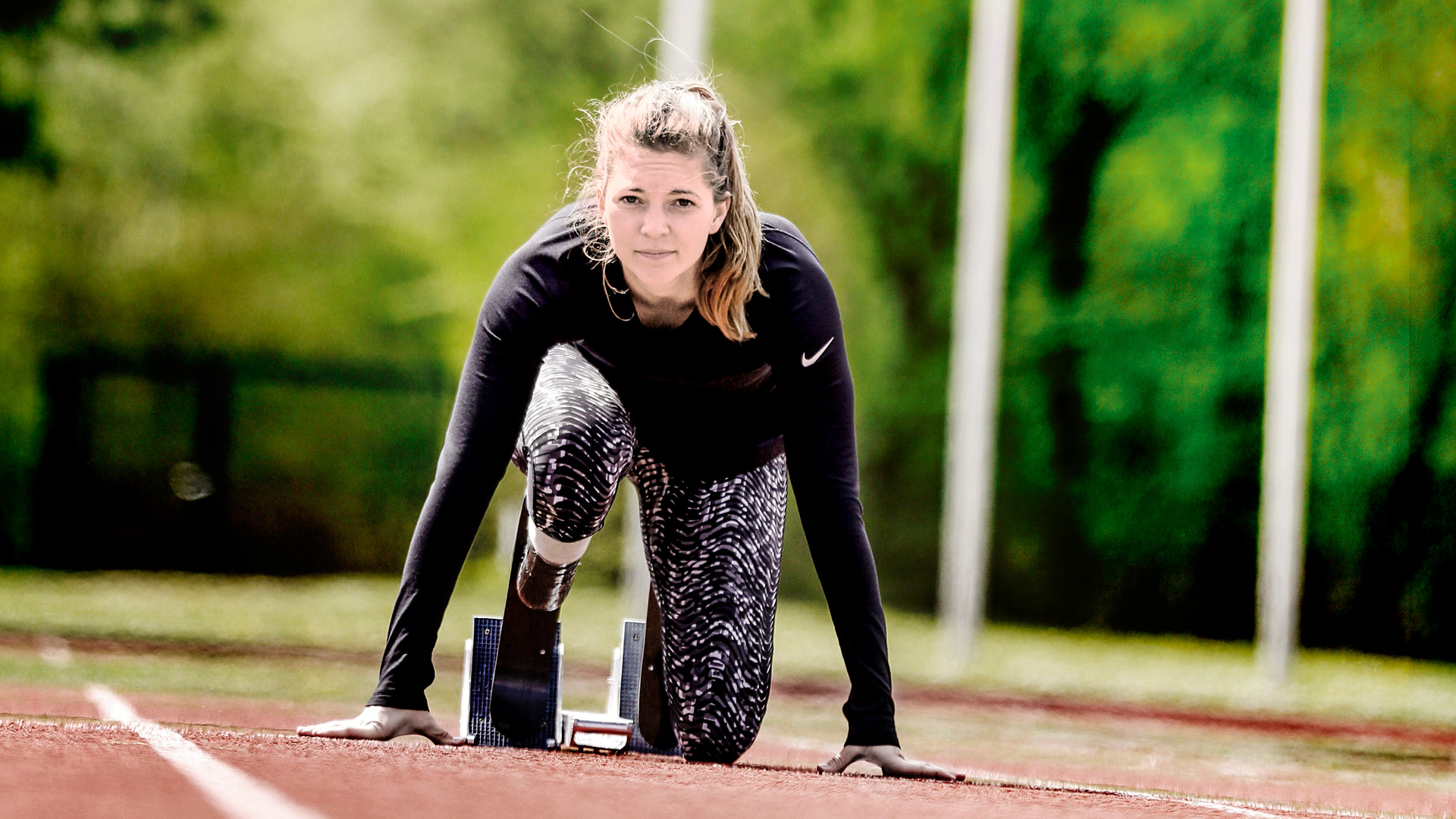 Eine beinamputierte Frau kniet auf einem Sportplatz in einem Startblock und trägt dabei eine Sportprothese von Ottobock.