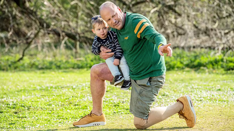 Auf dem Bild sieht man einen Mann, Axel. Er kniet draußen auf dem Boden, das andere Bein ist angewinkelt. Auf dem Bein sitzt ein kleines Kind, Axel zeigt mit dem Arm in eine Richtung in die beide schauen. Er trägt eine Beinprothese, das Genium Kniegelenk.