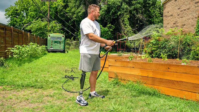 A C-Leg user is standing watering a flower bed with a hose pipe.