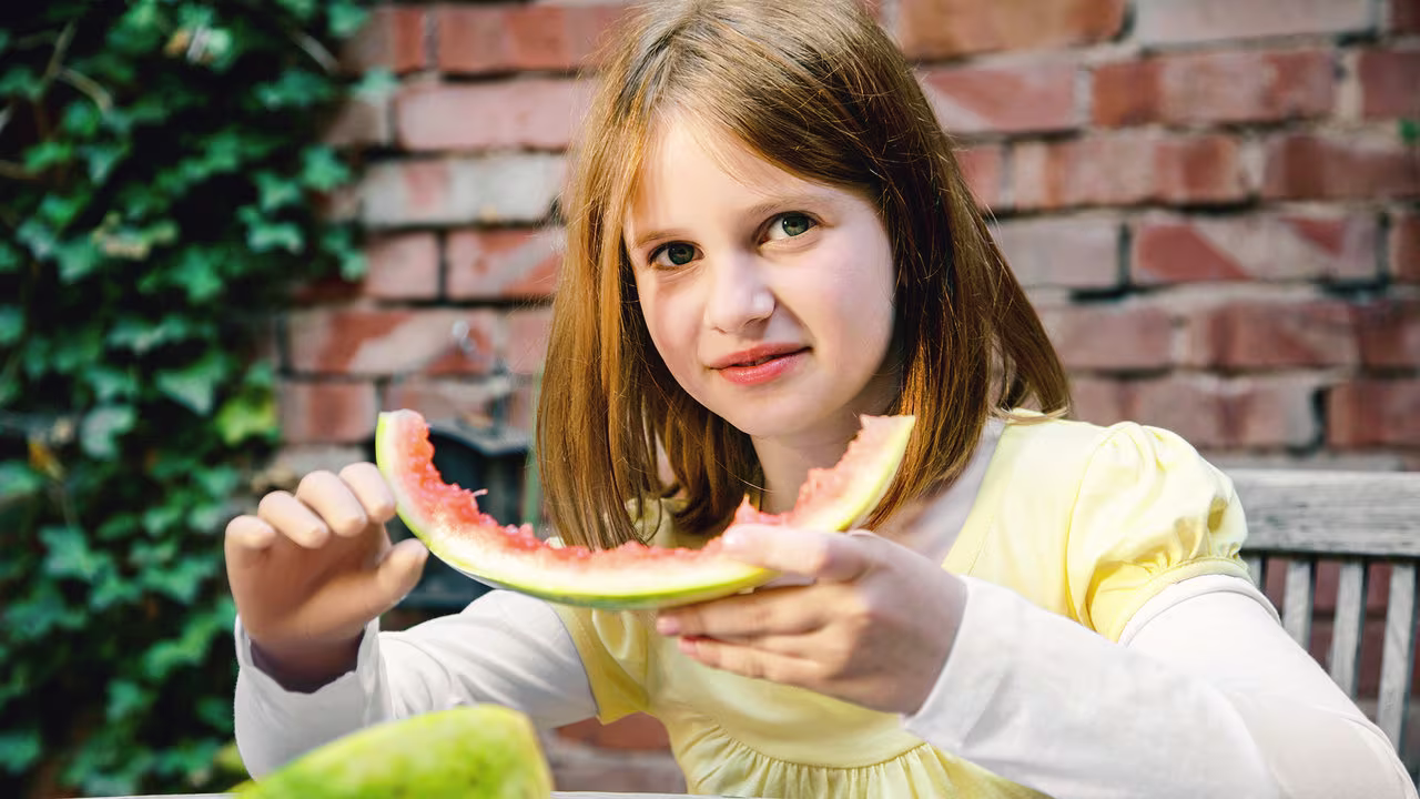 Eine junge Armprothesenträgerin isst eine Melone an einem Tisch. Sie trägt die Elektrohand 2000 von Ottobock. 