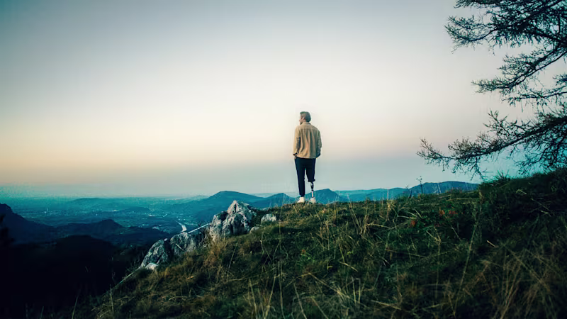 Un uomo con una protesi Ottobock è in piedi su una montagna con le spalle rivolte alla telecamera e guarda l'alba all'orizzonte.
