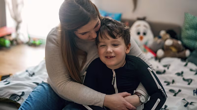 Ein kleiner Junge trägt den Exopulse Mollii Suit von Ottobock. Er sitzt mit seiner Mutter auf seinem Kinderbett. Sie umarmt ihn liebevoll von hinten.