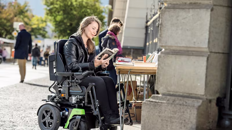 Rollstuhlfahrerin Kristina sitzt im Ottobock Juvo B4 an einem Bücherstand an der Straße und liest in einem Buch.