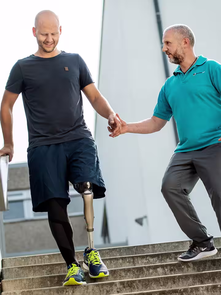 A man with a Kenevo from Ottobock walking down a set of outside stairs while being supported by his physiotherapist.