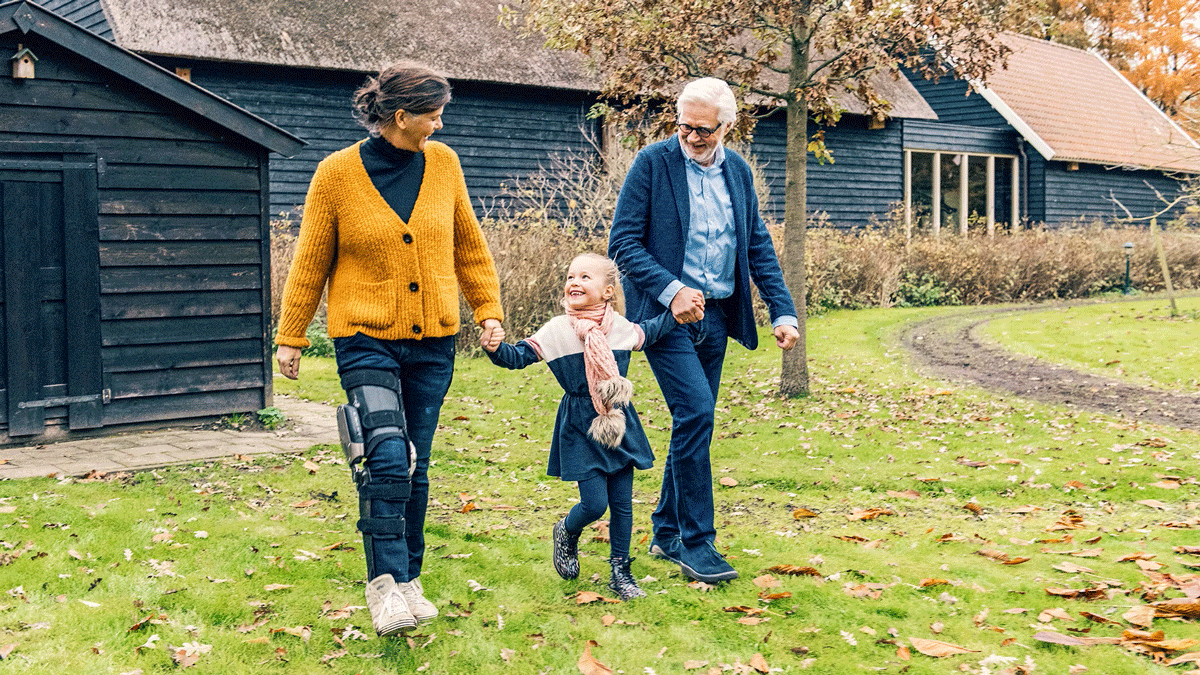 A man and woman walk hand in hand with their granddaughter, smiling. The woman is wearing an Ottobock C-Brace orthotic brace