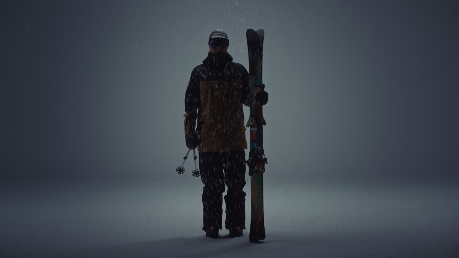 Skier in full ski clothing holding skis in his left hand and standing in front of a dark background.