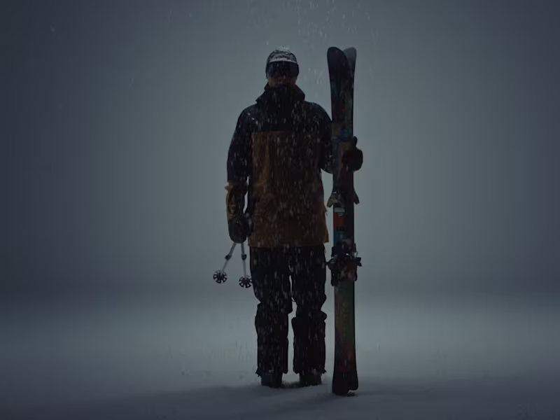 Skier in full ski clothing holding skis in his left hand and standing in front of a dark background.