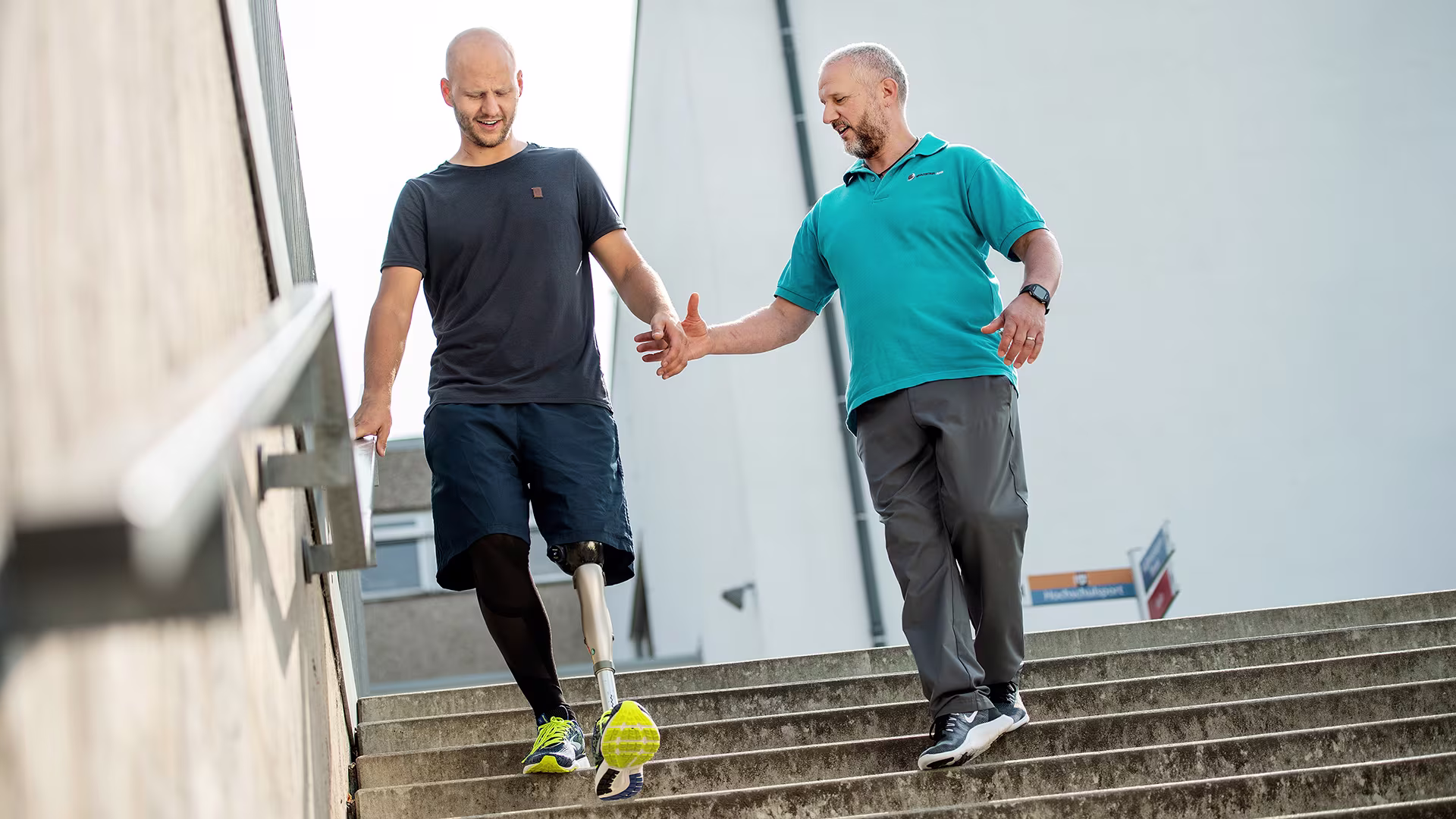 2 Männer gehen gemeinsam eine Treppe herunter. Einer der Männer trägt das mechatronische Kniegelenk Kenevo von Ottobock. Der andere Mann streckt unterstützend seine Hand zu dem Amputierten aus.