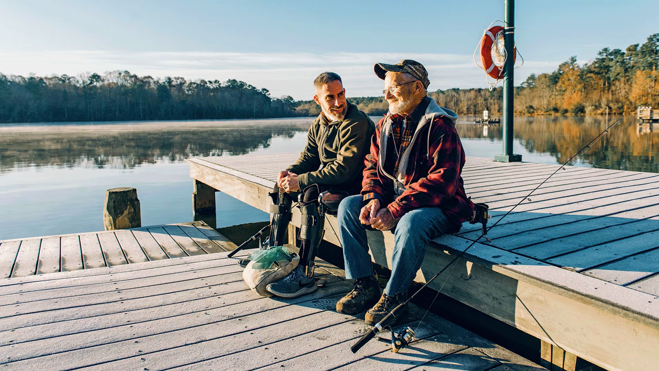 Two men sit on a dock talking and smiling with fishing gear nearby one of them is wearing an Ottobock C-Brace orthotic brace
