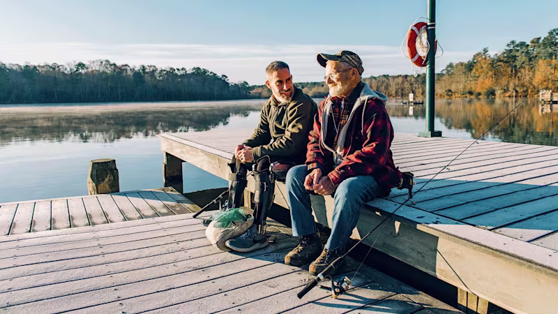 Two men sit on a dock talking and smiling with fishing gear nearby one of them is wearing an Ottobock C-Brace orthotic brace