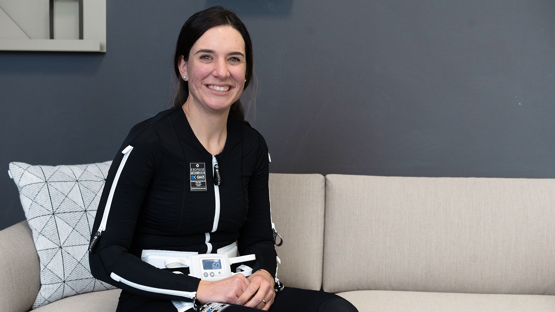 Eine dunkelhaarige Frau trägt den Exopulse Mollii Suit von Ottobock. Sie sitzt auf einem beigen Sofa und lächelt in die Kamera.