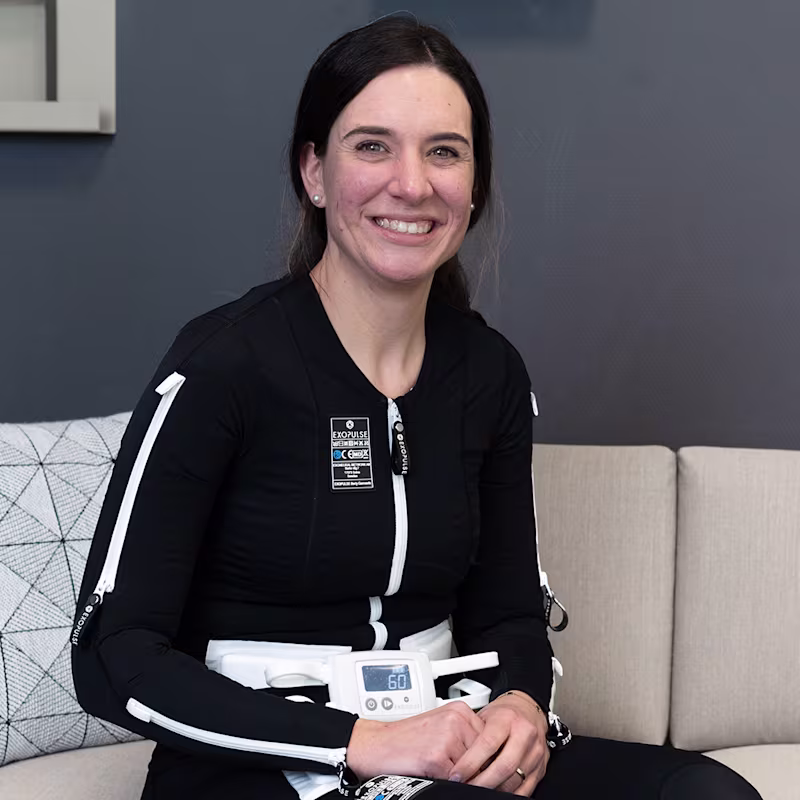 Eine dunkelhaarige Frau trägt den Exopulse Mollii Suit von Ottobock. Sie sitzt auf einem beigen Sofa und lächelt in die Kamera.