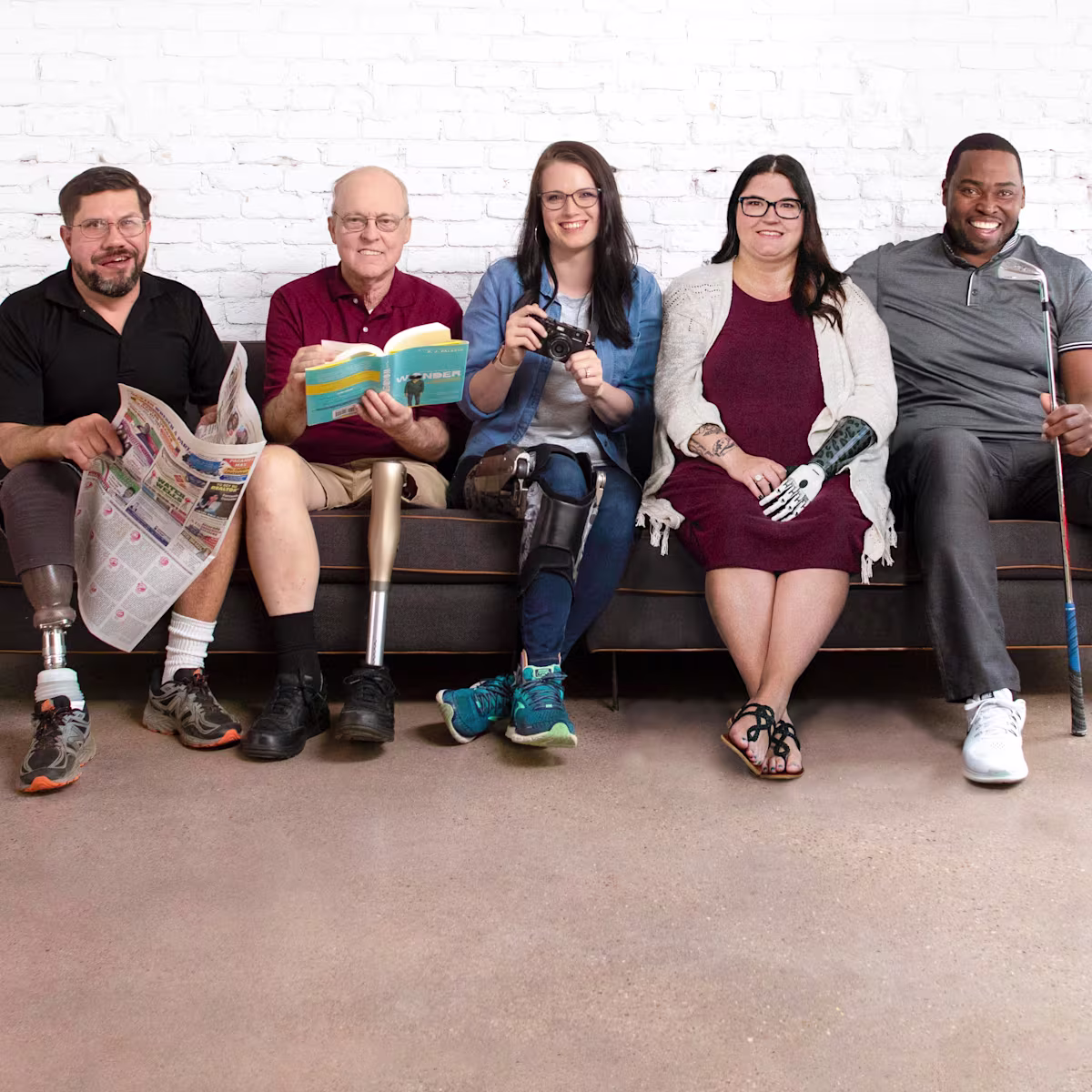 Five people with limb loss and limb difference happily sit on a couch together in front of a white brick wall.