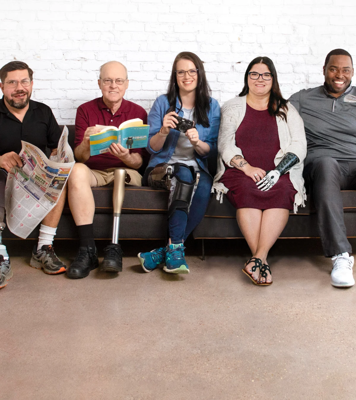 Five people with limb loss and limb difference happily sit on a couch together in front of a white brick wall. 