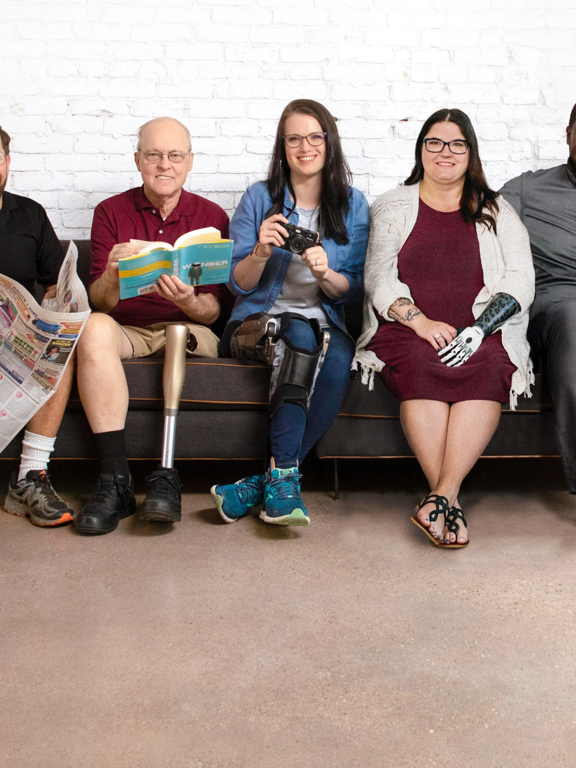 Five people with limb loss and limb difference happily sit on a couch together in front of a white brick wall. 