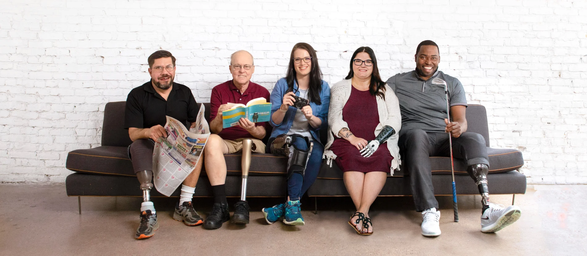 Five people with limb loss and limb difference happily sit on a couch together in front of a white brick wall. 