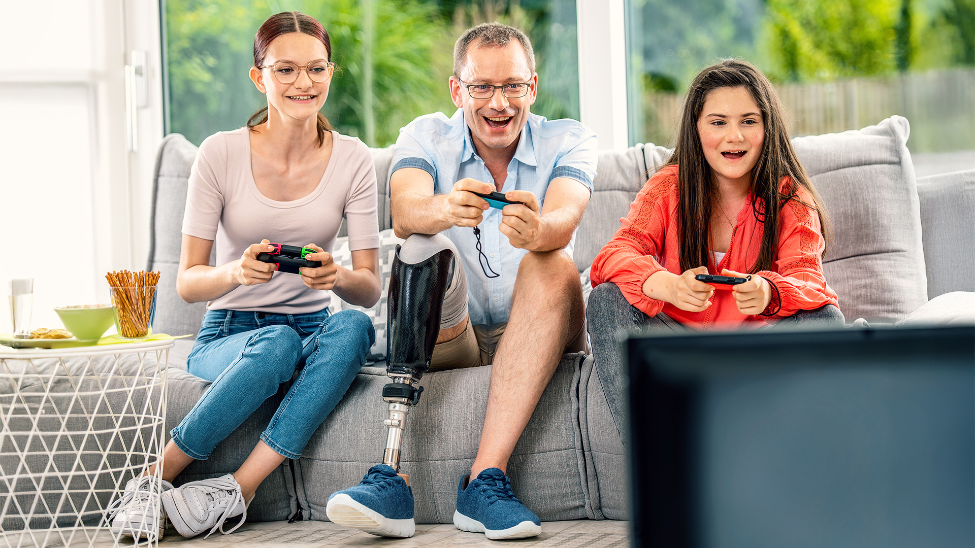 Two girls and a man with a prosthetic leg sit next to each other on the sofa with joysticks in their hands.