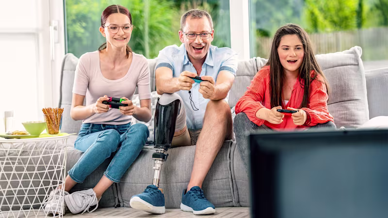 Two girls and a man with a prosthetic leg sit next to each other on the sofa with joysticks in their hands.