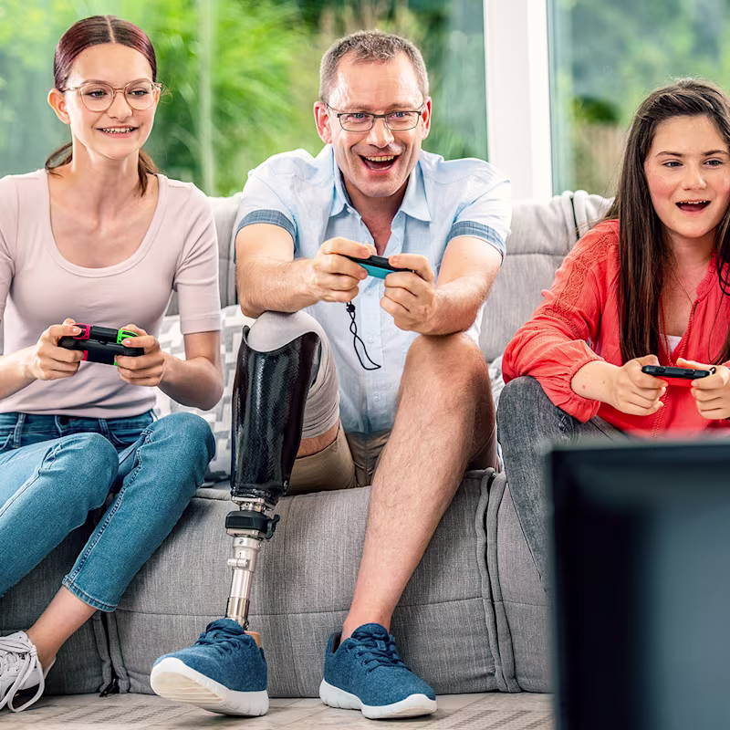 Two girls and a man with a prosthetic leg sit next to each other on the sofa with joysticks in their hands.