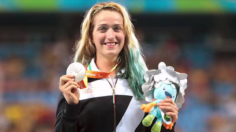 A young woman with blond and blue hair holding up a silver medal and a stuffed animal, smiling.