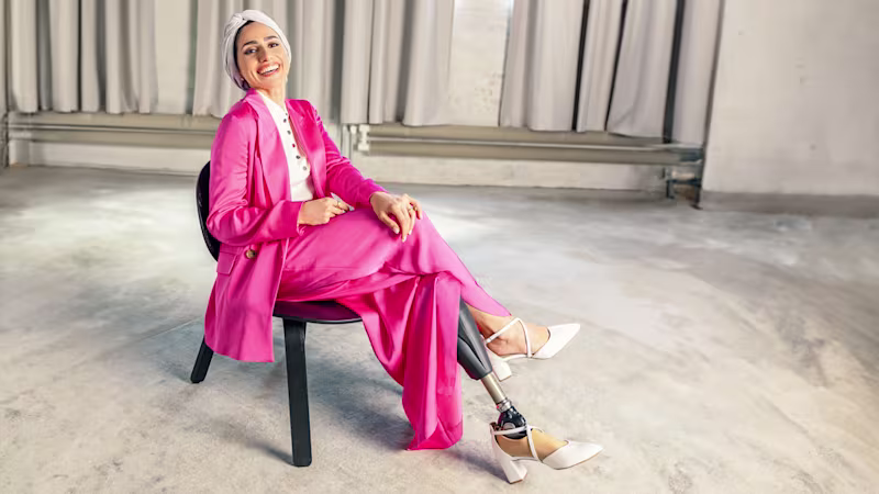 A young woman with a prosthetic leg sitting on a chair and laughing, wearing a pink suit, a turban, and high heels.
