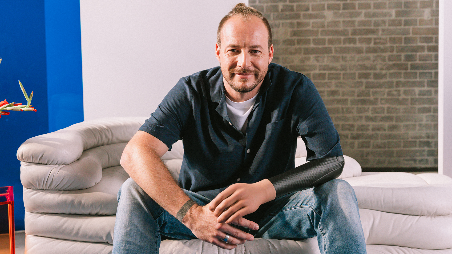 A man with a Dynamic Arm elbow prosthesis sitting on a white sofa in front of a brick wall