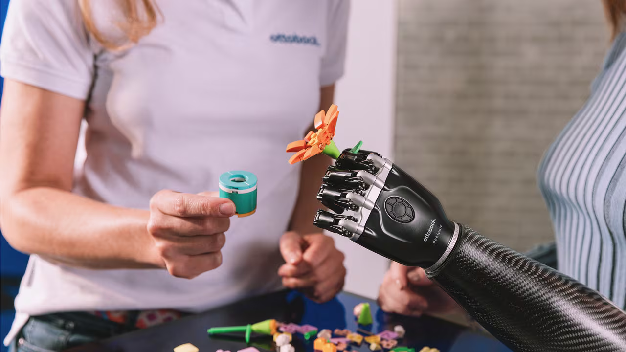 Eine Armprothesenträgerin sitzt an einem Tisch und hält eine Plastikblume in der Hand. Sie trägt die bebionic Hand von Ottobock. 