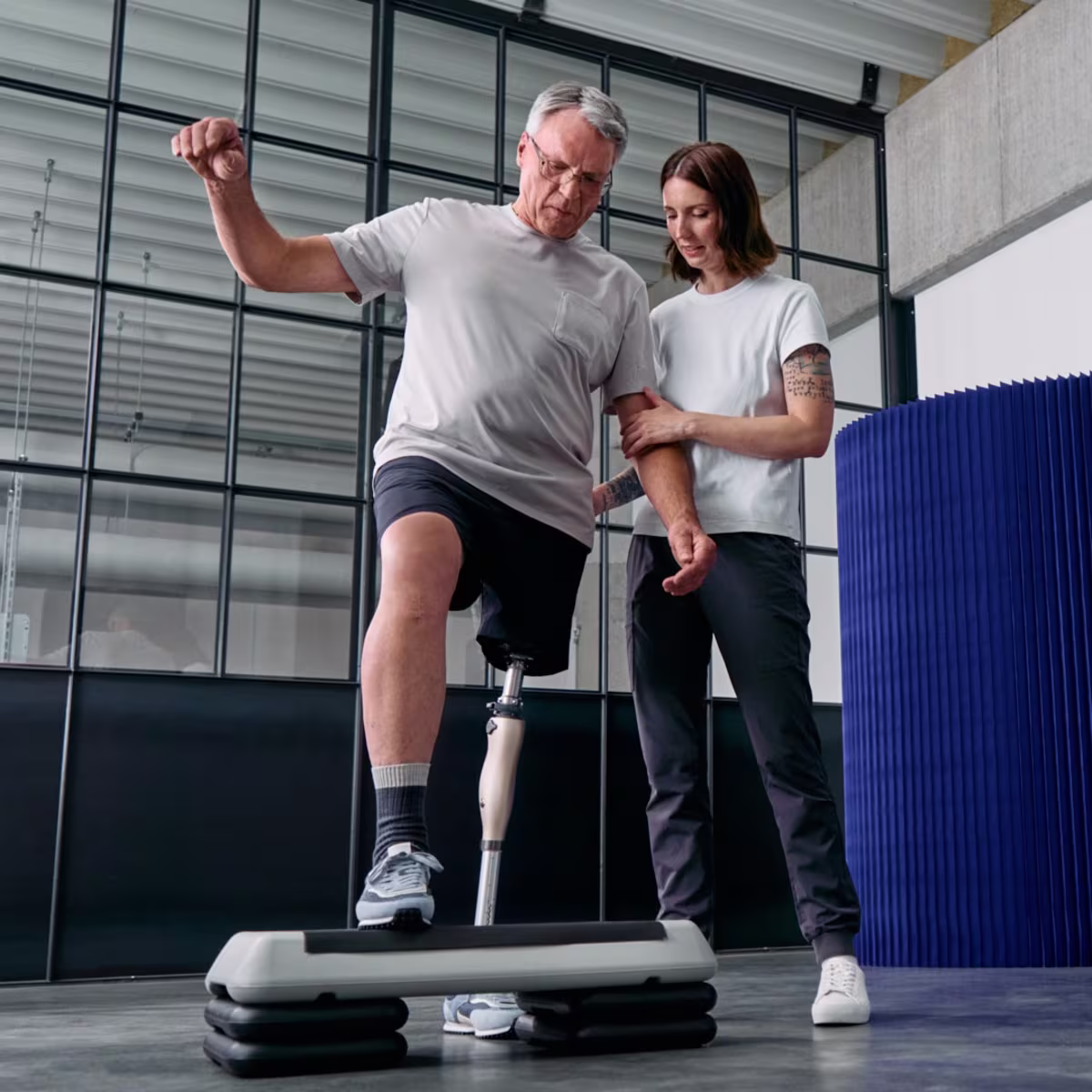 Ein Mann und seine Physiotherapeutin stehen in einer Sporthalle vor einem Stepbrett. Der Mann trägt das Kenevo Interimsprothesengelenk von Ottobock und stellt sich auf das Stepbrett.