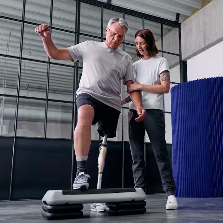 Ein Mann und seine Physiotherapeutin stehen in einer Sporthalle vor einem Stepbrett. Der Mann trägt das Kenevo Interimsprothesengelenk von Ottobock und stellt sich auf das Stepbrett.