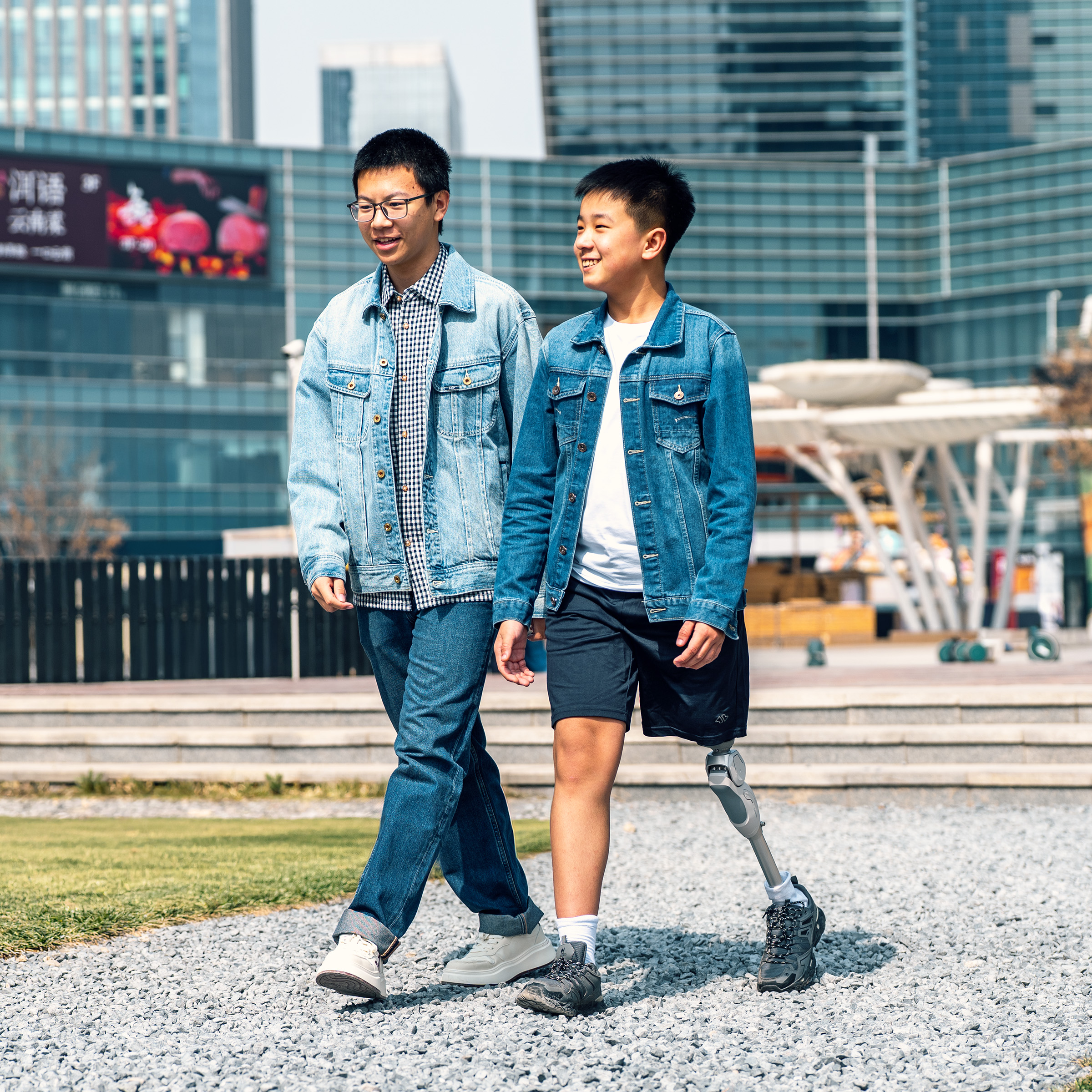 Two boys walk side by side through a mall courtyard, one using a leg prosthesis.