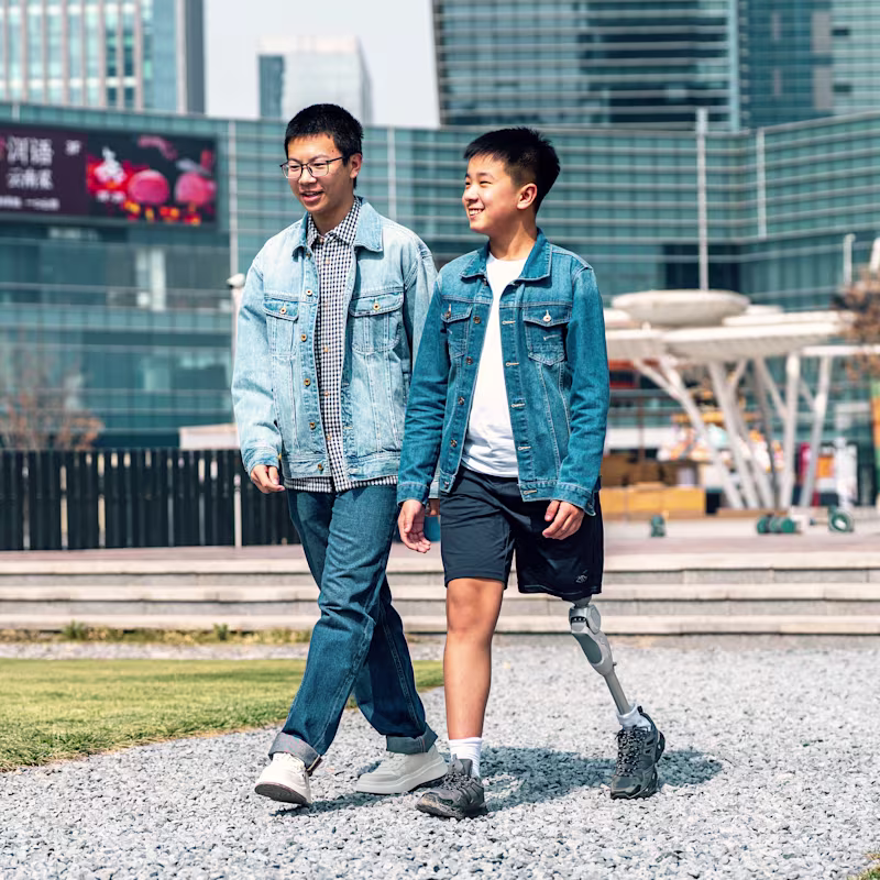 Two boys walk side by side through a mall courtyard, one using a leg prosthesis.