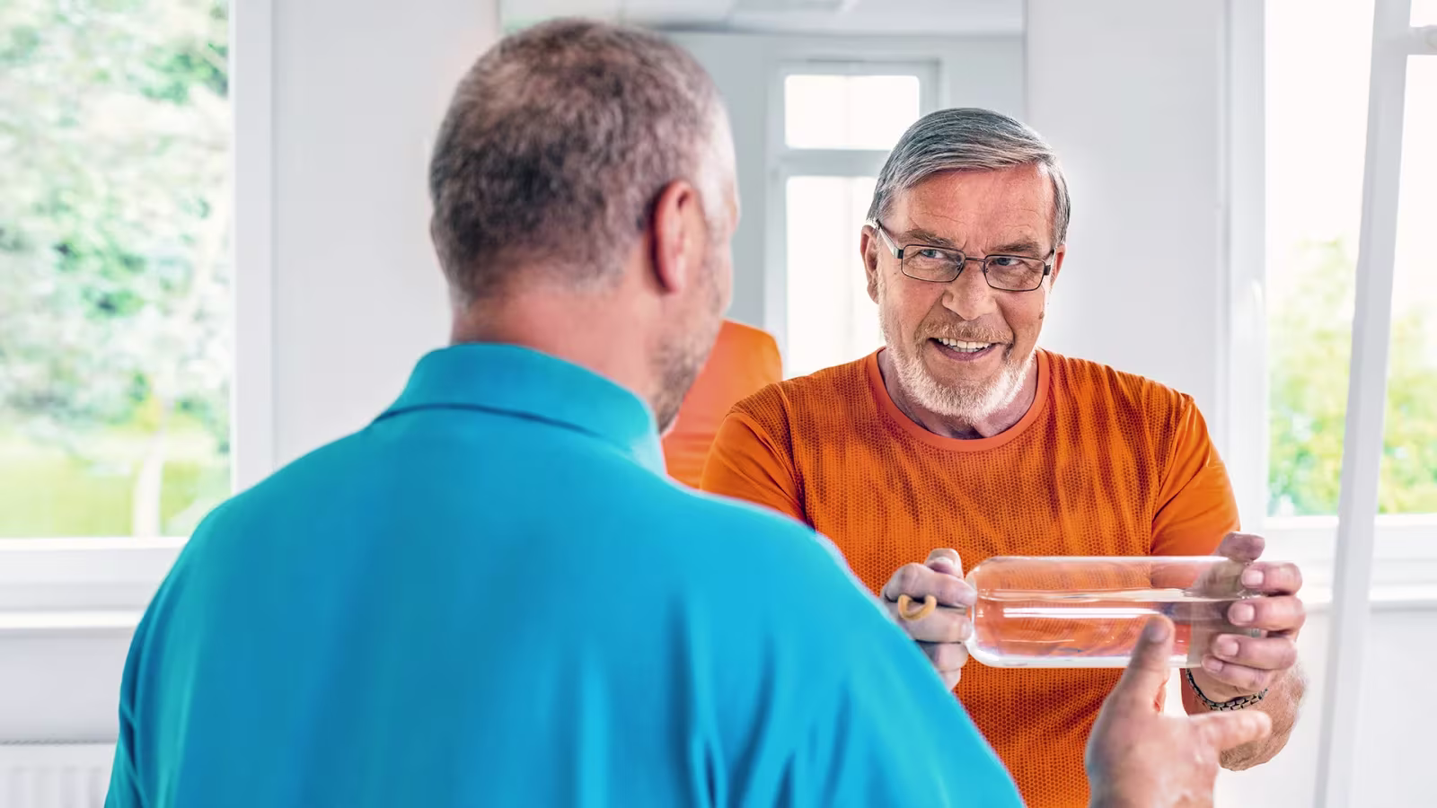 Two people, one in a blue shirt and one in an orange shirt, interacting with a transparent rectangular object indoors, with windows and greenery in the background.