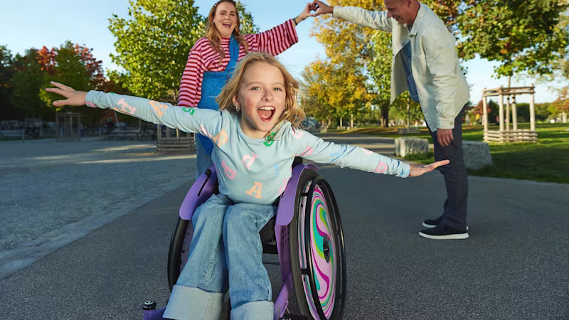 Ein weiblich gelesenes Kind sitzt im Kiddo Rollstuhl von Ottobock. Sie streckt die Arme zu beiden Seiten und sieht dabei sehr fröhlich aus. Hinter ihr stehen eine weiblich und eine männlich gelesene Person, die ihre Arme zu einem Tunnel formen.