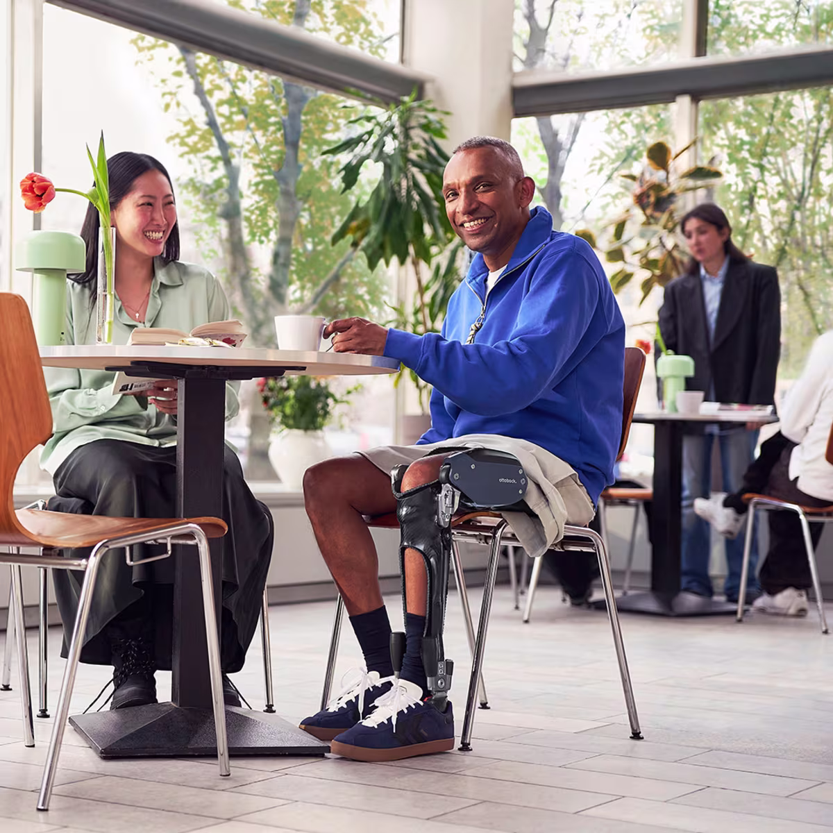 A man and a woman sit smiling at a cafe table. The man is wearing an Ottobock C-Brace orthotic brace