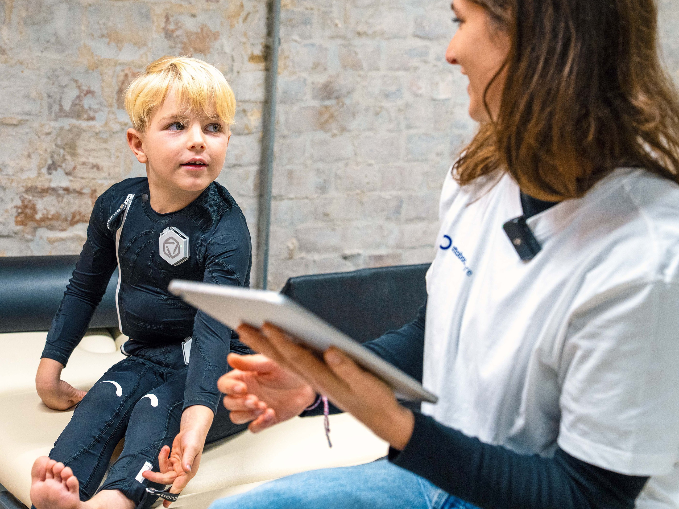 Young boy wearing Exopulse neuromodulation suit sitting on examination table, therapist reviewing treatment data on a tablet during pediatric rehabilitation session