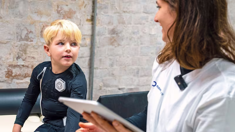 Young boy wearing Exopulse neuromodulation suit sitting on examination table, therapist reviewing treatment data on a tablet during pediatric rehabilitation session