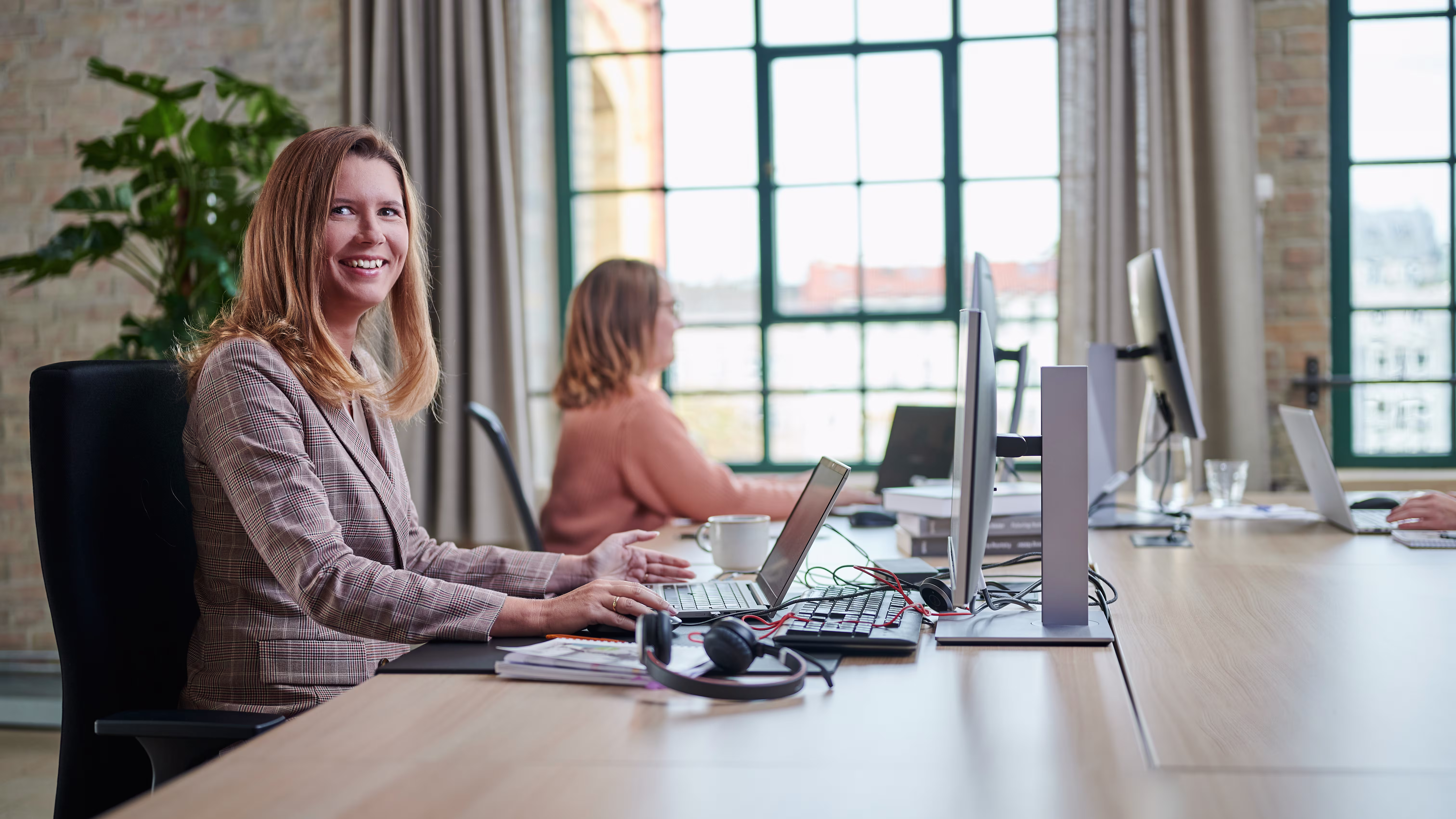 2 Frauen sitzen nebeneinander im Büro am Computer. Eine der beiden Frauen schaut auf den Bildschirm, die andere lächelt freundlich in die Kamera.