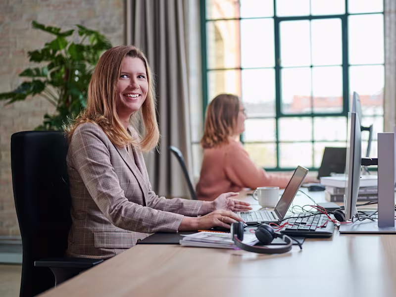 2 Frauen sitzen nebeneinander im Büro am Computer. Eine der beiden Frauen schaut auf den Bildschirm, die andere lächelt freundlich in die Kamera.