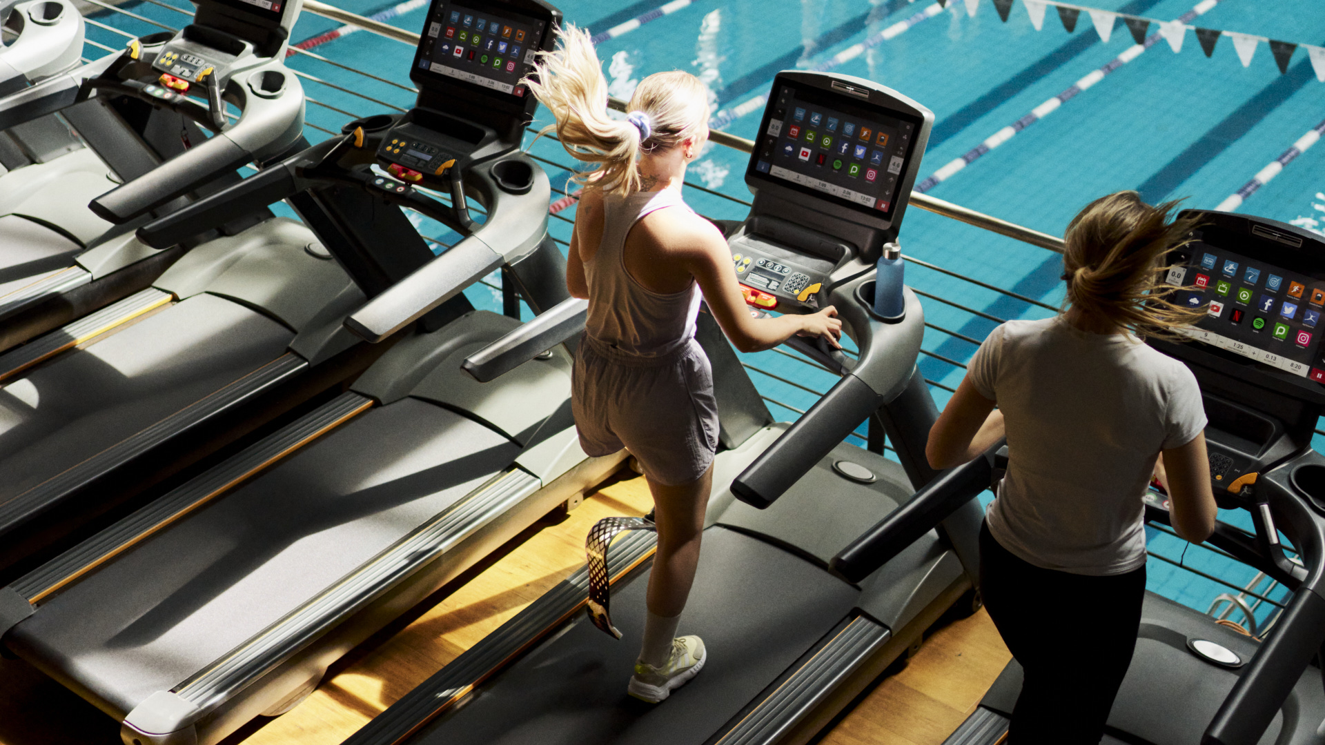 Eine junge Frau läuft in einem Fitnessstudio auf dem Laufband. Das Bild zeigt sie von hinten. Im Hintergrund ist ein Pool zu sehen. Die Frau trägt eine Sportprothese von Ottobock,