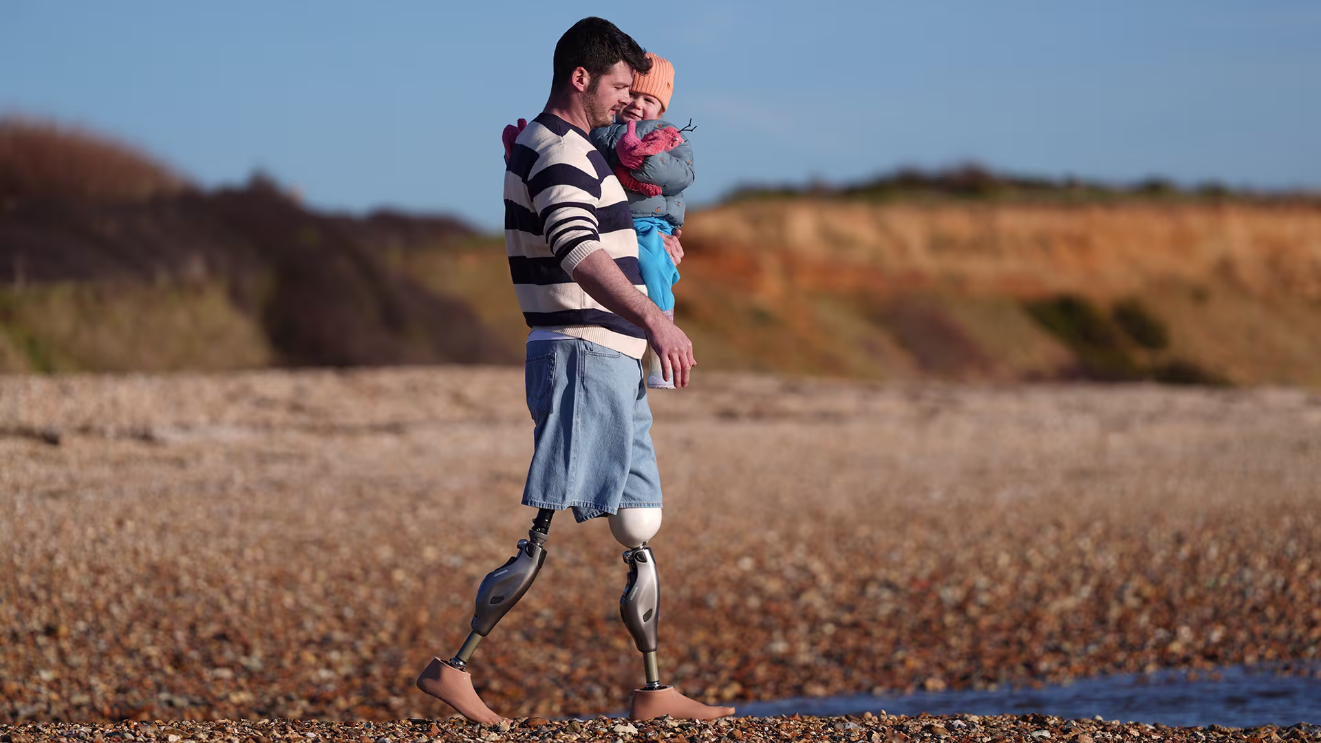 Veteran and bilateral Genium X4 MPK user Dave Henson walks across a pebble beach holding his daughter.