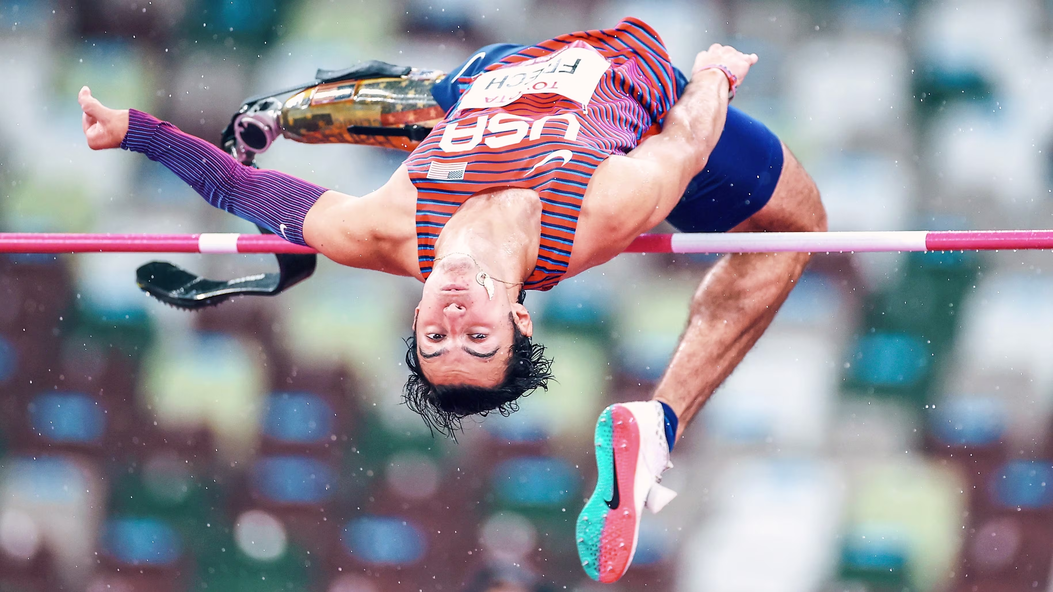 A young dark-haired man with a prosthetic leg high jumping with his body upside down.