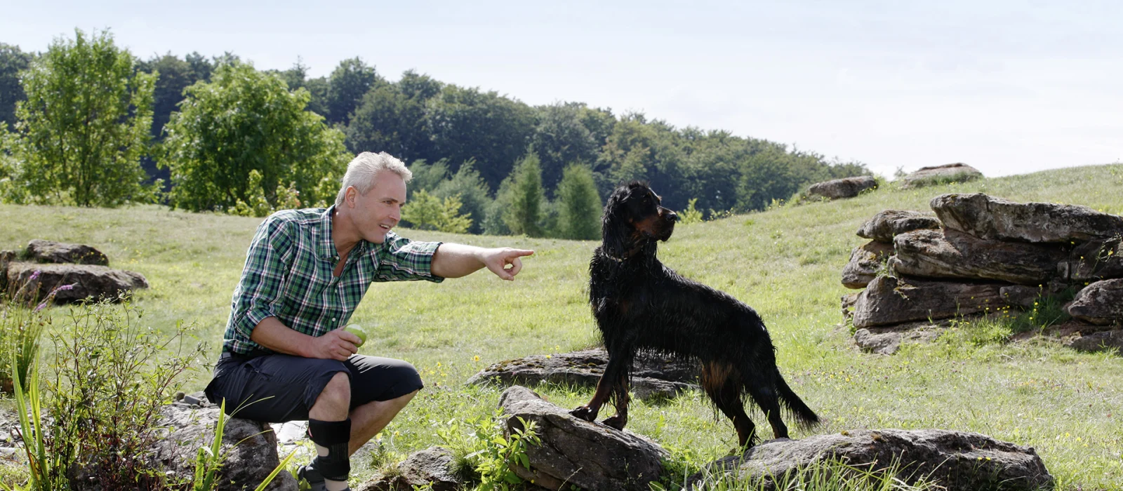 A man sitting on a rock outdoors directs his dog's attention as he points to something in the distance. The man is wearing an Ottobock brace on his ankle.