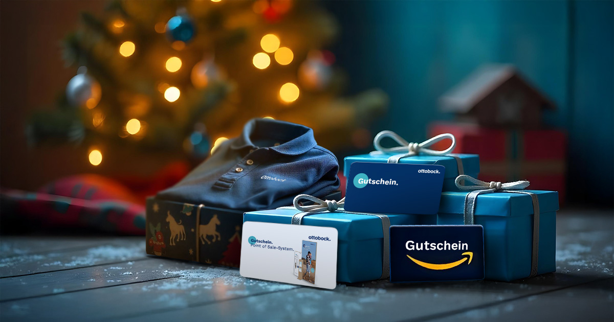 Ein festlich geschmückter Weihnachtsbaum im Hintergrund mit warmen Lichtern. Im Vordergrund sind blaue, weihnachtlich verpackte Geschenke zu sehen, darunter Gutscheine von Ottobock und Amazon sowie ein dunkelblaues Polo-Shirt mit Ottobock-Logo. Die Szene ist auf einer Holzoberfläche mit leichtem Kunstschnee gestaltet, was eine gemütliche Weihnachtsatmosphäre schafft.