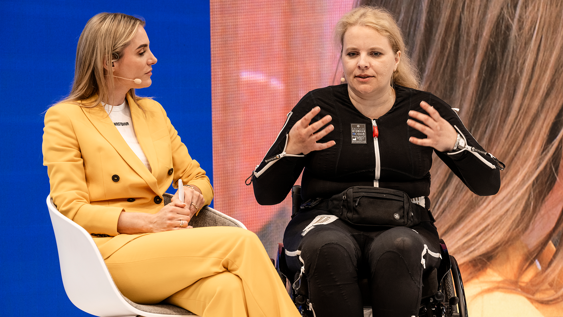 TV-Moderatorin Anna Kraft und eine Exopulse Mollii Suit Anwenderin im Rollstuhl auf der Show-Bühne von Ottobock auf der OTWorld 2024 in Leipzig.